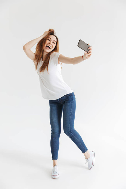 Full length image of Playful woman in casual clothes making selfie on smartphone while touching hair over grey background - Foto, imagen