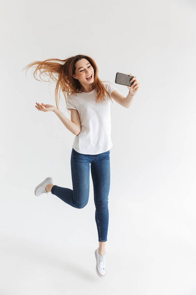 Full length image of Playful woman in casual clothes making selfie on smartphone and jumping over grey background - Foto, Bild