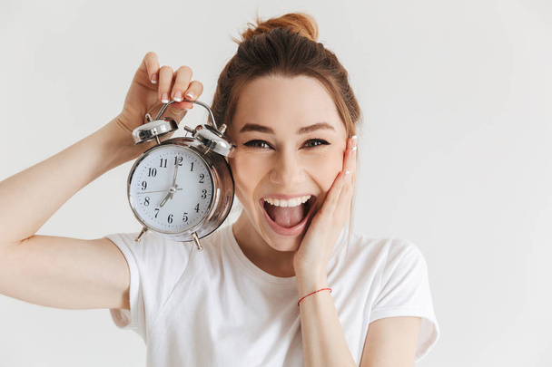 Happy laughing woman in casual clothes holding alarm clock and looking at the camera with arm on cheek over grey background - Photo, image