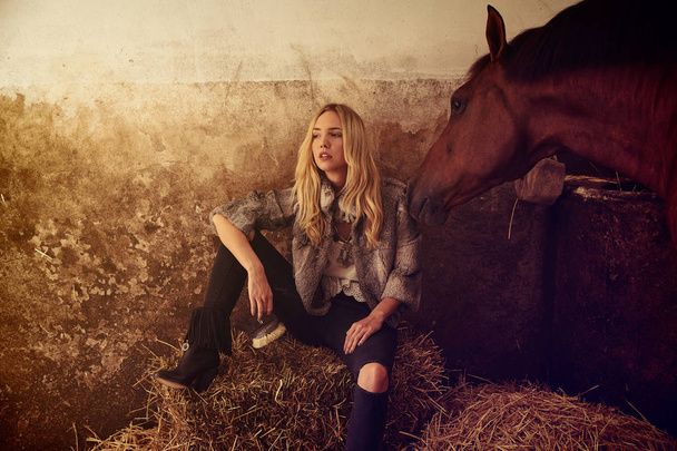 Blond woman on an indoor stable with a brown horse on straw bale - Foto, Imagem