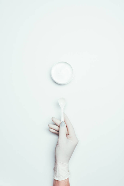 cropped image of woman in latex glove holding spoon near container with clay mask isolated on white surface  - Foto, afbeelding