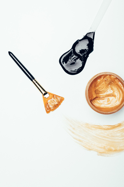 Konteyner, fırça, leke ve kaşık altın maskesi ile siyah maske beyaz yüzey üzerinde izole üstten görünüm  - Fotoğraf, Görsel