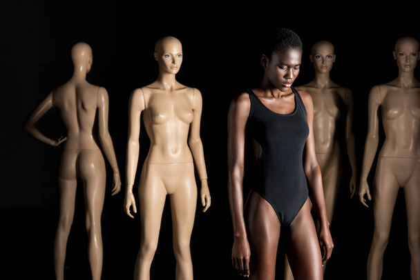 young african american woman in swimsuit standing in front of mannequins and looking down on black   - Zdjęcie, obraz