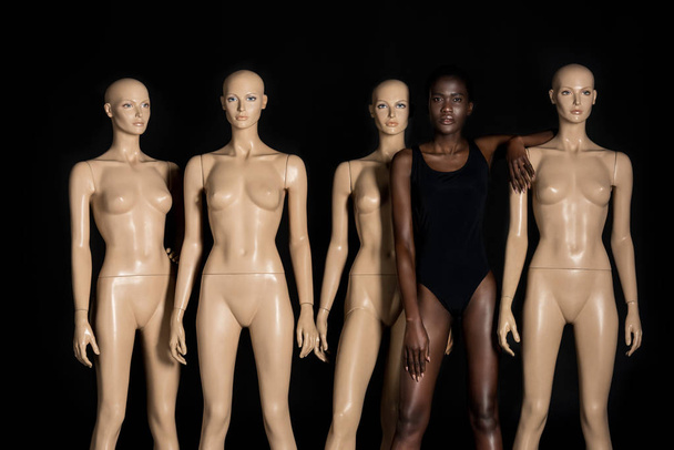 beautiful young african american woman in swimsuit standing between mannequins and looking at camera on black  - Фото, зображення