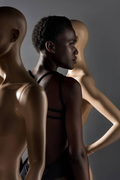 side view of beautiful african american girl looking away while standing between dummies on black - Φωτογραφία, εικόνα