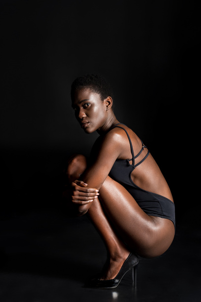 vista lateral de la hermosa chica afroamericana en traje de cuerpo agachado y mirando a la cámara en negro
 - Foto, Imagen