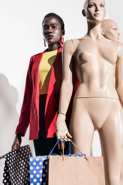 low angle view of young african american woman looking at camera while standing near dummies with shopping bags on white - Photo, image