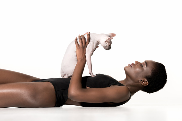 side view of beautiful young african american woman in swimsuit lying and holding sphynx cat isolated on white - Photo, Image