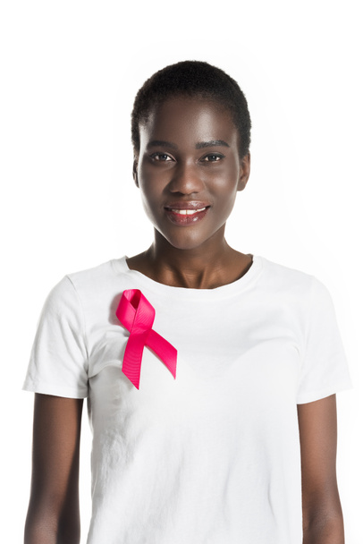 genç Afro-Amerikan kadın t-shirt kameraya gülümseyen üzerinde pembe kurdele ile izole beyaz, meme kanseri kavramı üzerinde - Fotoğraf, Görsel