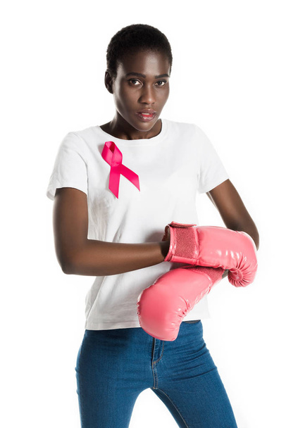 fiatal afrikai-amerikai nő, rózsaszín szalag, boksz-kesztyűket visel, és látszó-on fényképezőgép elszigetelt fehér, mell rák fogalma - Fotó, kép