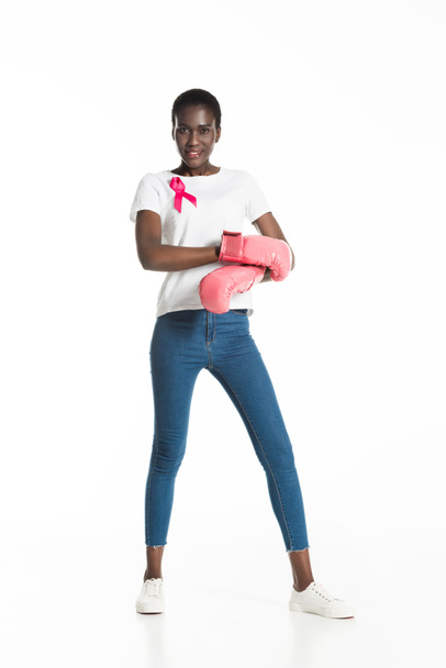 full length view of young woman with pink ribbon wearing boxing gloves and smiling at camera isolated on white, breast cancer concept - Photo, Image