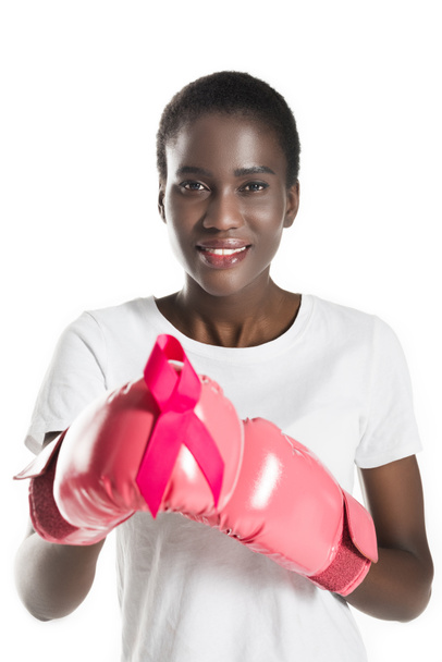 nuori afrikkalainen amerikkalainen nainen nyrkkeilyhanskat tilalla vaaleanpunainen nauha ja hymyillen kameran eristetty valkoinen, rintasyöpä käsite
 - Valokuva, kuva