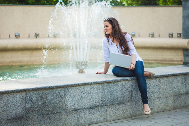 beautiful business girl brunette sitting with laptop at a fountain in the street. - Zdjęcie, obraz