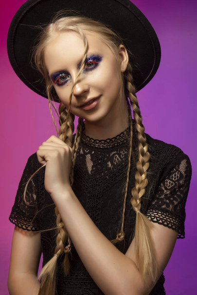 A beautiful smiling blonde teenage girl wearing a black gothic dress and a black hat. Conceptual makeup, red eyelashes and sequins. Healthy smooth skin. Pink and violet background. Copy space. - Photo, image