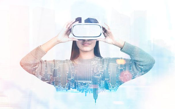 Portrait of a young woman with long dark hair wearing a gray sweater and a pair of VR glasses. A cityscape background. Concept of the future technology. Toned image double exposure - Foto, afbeelding