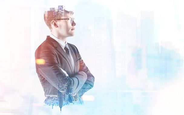 Side view of a confident young businessman wearing a suit and glasses and standing with crossed arms looking into the distance. A blurred foggy cityscape. Toned image double exposure mock up - Photo, Image