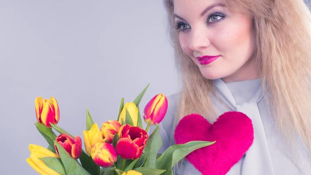 International womens or valentines day. Attractive grateful woman blonde hair holding tulips bunch and red heart sign. On grey - Foto, afbeelding