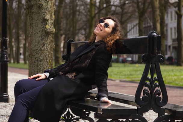 A girl in a black coat with long curly hair enjoying the sun. Attractive young woman in black glasses sitting in a spring park on a bench. - Valokuva, kuva