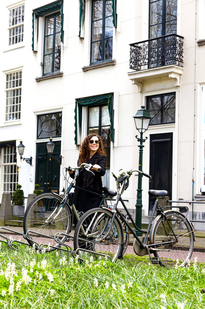 A young girl stands on a white beautiful house background with a bicycle. Woman in sunglasses parking her vintage bike on the street in old town. - Фото, изображение