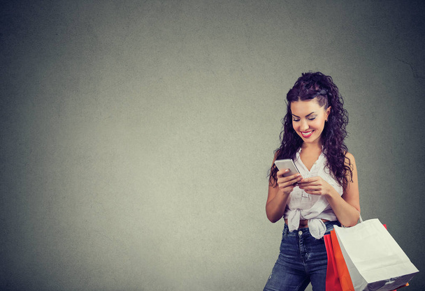 Opgewonden trendy meisje met papieren tassen met smartphone om te winkelen in Internet glimlachend op grijze achtergrond.  - Foto, afbeelding