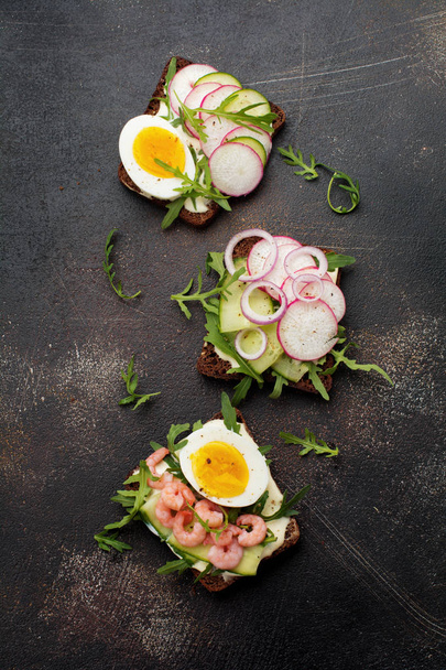 Open sandwiches on dark rye bread with eggs, shrimps, radishes, cucumber, cream cheese and arugula for breakfast on old dark background. Smorrebrod Traditional dish of Danish cuisine. Top view. - Photo, image
