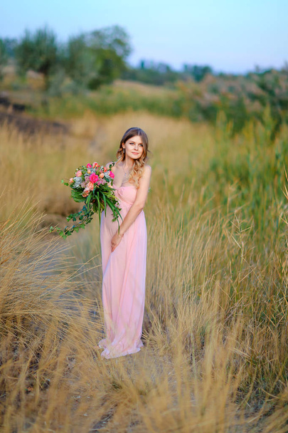 Zwangere vrouw draagt roze jurk en boeket bloemen staan in de steppe achtergrond. - Foto, afbeelding