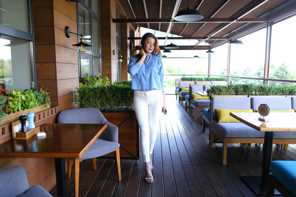 Młody Fotografia model stojący w kawiarni i utrzymanie smartphone, noszenie dżinsów koszulę i białe spodnie. - Zdjęcie, obraz
