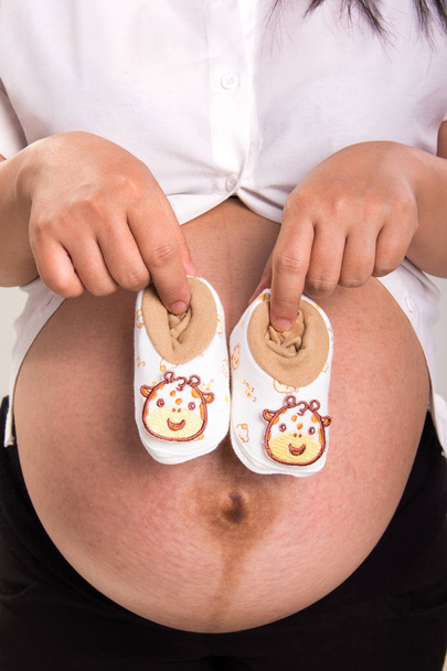 Χέρια των πρόσωπων που κρατά ένα ζευγάρι παπούτσια μωρών με χαριτωμένο ζώο σχεδιασμό, τοποθετείται μπροστά από την κοιλιά της - Φωτογραφία, εικόνα