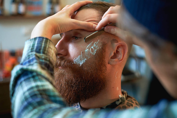Nahaufnahme einer Friseurarbeit für einen attraktiven jungen blonden Mann beim Friseur. er stylt seinen Bart, rasiert sich mit Rasiermesser - Foto, Bild