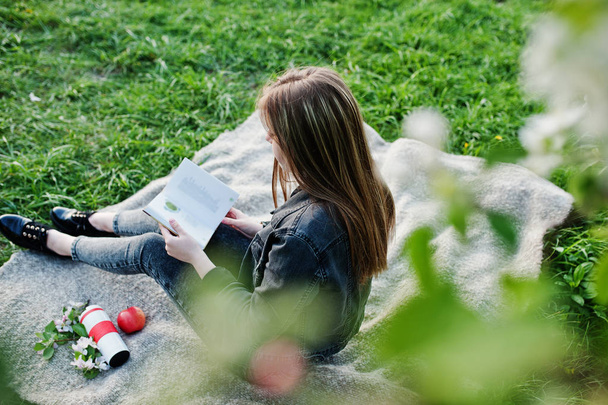 Young brunette girl at jeans sitting on plaid against spring blossom tree and read the book. - Фото, зображення