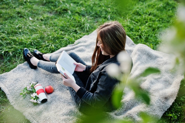 Young brunette girl at jeans sitting on plaid against spring blossom tree and read the book. - Fotó, kép