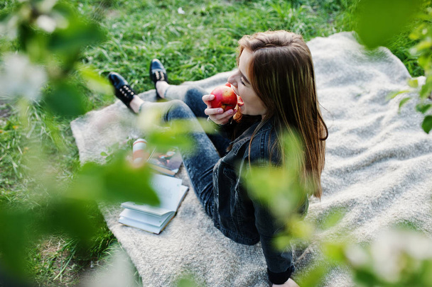 Young brunette girl at jeans sitting on plaid against spring blossom tree and read the book and eat apple. - 写真・画像