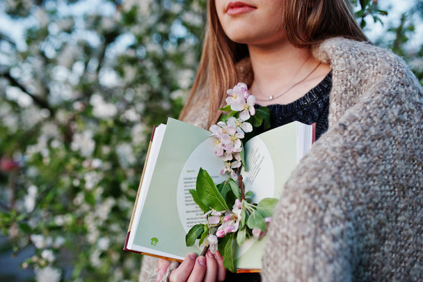 Young brunette girl on plaid against spring blossom tree and holding book at hands. - Foto, Imagen