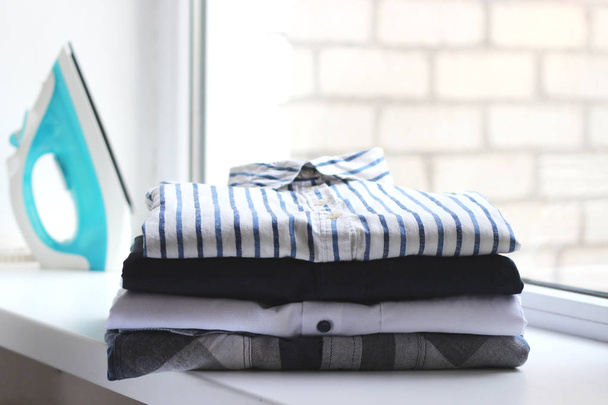 Una pila de ropa y una plancha sobre un fondo claro. minimalismo, planchado de ropa
. - Foto, Imagen