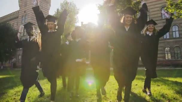 Více etnických absolventů v černé šaty a čepice vesele běží s jejich diplomů v ruce před jejich univerzity za slunečného dne. Venkovní - Záběry, video