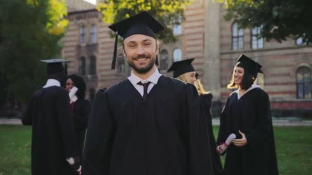 Portrait of the handsome young graduate in black traditional gown and cap posing to the camera and doing YES gesture. Graduates on the background. Outdoor - Footage, Video