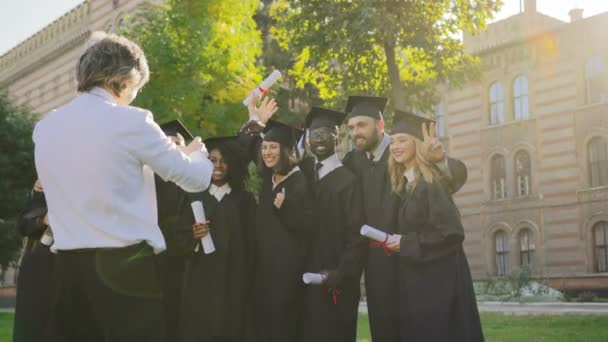Vista posterior del hombre de pelo gris tomando un retrato fotográfico en el teléfono inteligente negro de los graduados multiétnicos en ropa negra tradicional y gorras con diplomas en las manos. Al aire libre
 - Metraje, vídeo