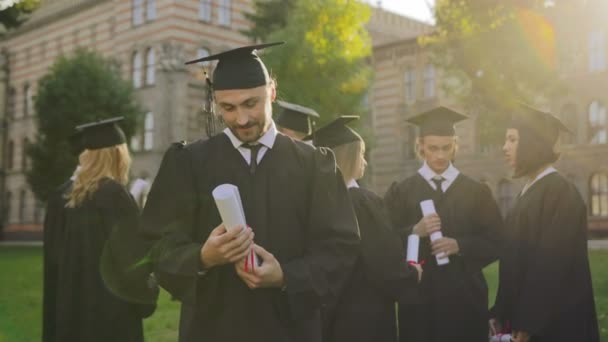 portrait shot of the attractive young man in graduation gown and cap looking at the diloma in his hands and than to the camera. His friends on the background. Outdoor - Footage, Video