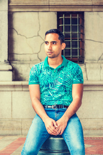 Young Hispanic American Man thinking outside in New York, wearing green patterned Polo shirt, sitting on vintage street, looking away, relaxing - Photo, Image