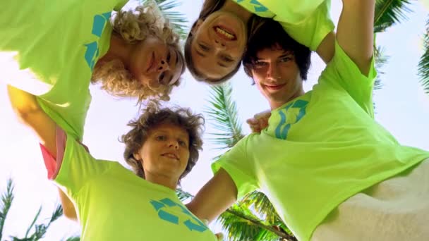 Čtyři mladí dobrovolníci v zelených tričkách s obrázkem recyklaci tvořící hloučcích pod palmami na pobřeží oceánu pláže, po vyčištění pláže od plastové úlomky. Dobrovolnictví a - Záběry, video