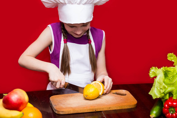 Szczęśliwa dziewczynka w jednolity kuchni cięcia owoców w kuchni. Szef kuchni dziecko. Proces gotowania - Zdjęcie, obraz