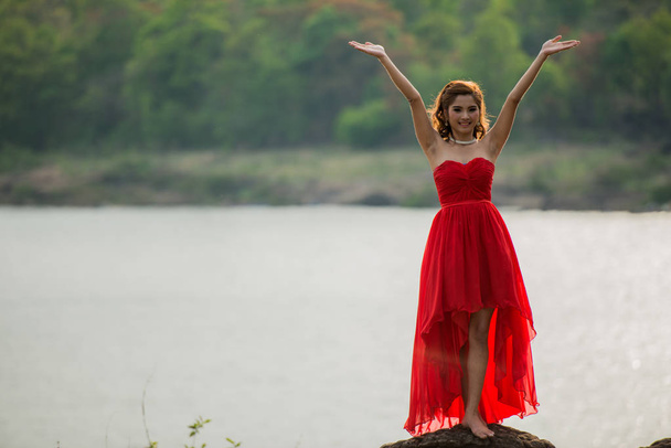 a bela menina com o cabelo longo no vestido fluttering vermelho custa no banco de um fluxo
 - Foto, Imagem