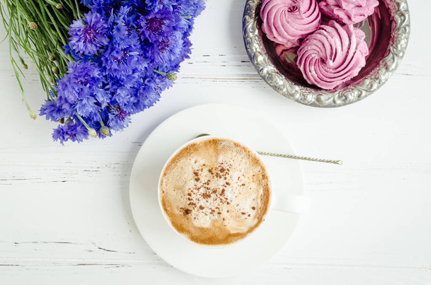 Pink zephyr marshmallow with cup of cappuccino coffee and bouquet of blue cornflowers on white wooden table. Top view. - Valokuva, kuva