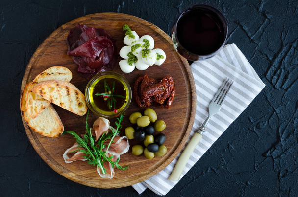 Italian antipasti wine snacks set. Mozzarella cheese, Mediterranean olives, Prosciutto di Parma, bresaola, sun-dried tomatoes and toasts on wooden board and red wine in glass. Spanish tapas. Top view. - Фото, изображение
