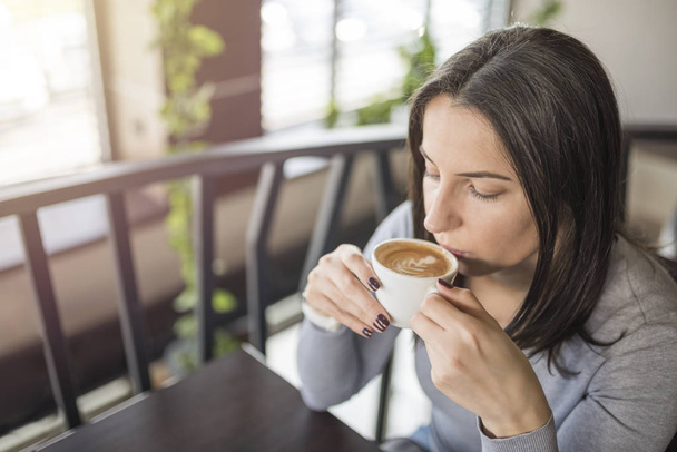 Young beautiful girl sitting in coffee shop and enjoying coffee. Young woman drinking coffee in restaurant. Female holding cup of coffee. Relax time. - Photo, image