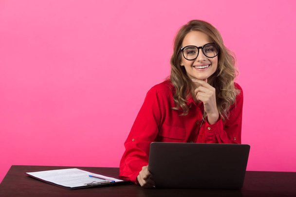 beautiful young girl in a red shirt and glasses near a laptop on a pink background - 写真・画像