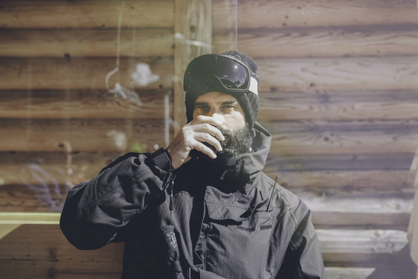 Bearded handsome snowboarded taking rest after ride session.Young man drinking cup of hot coffee on sunny terrace. Blurred background.Horizontal.Visual effects. - Foto, afbeelding