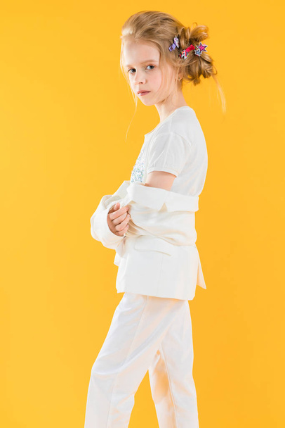 Een tienermeisje in witte kleren poserend op een gele achtergrond. Het meisje liet haar jas naar haar ellebogen zakken.. - Foto, afbeelding