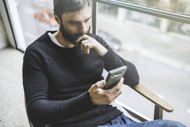Handsome bearded man using smartphone while resting in modern studio loft. Blurred background. - Фото, изображение