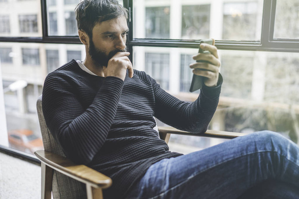 Young cute bearded man sitting home and using smartphone for surfing web internet. Man using gadget at modern apartment. Horizontal. Blurred background. - Foto, Bild